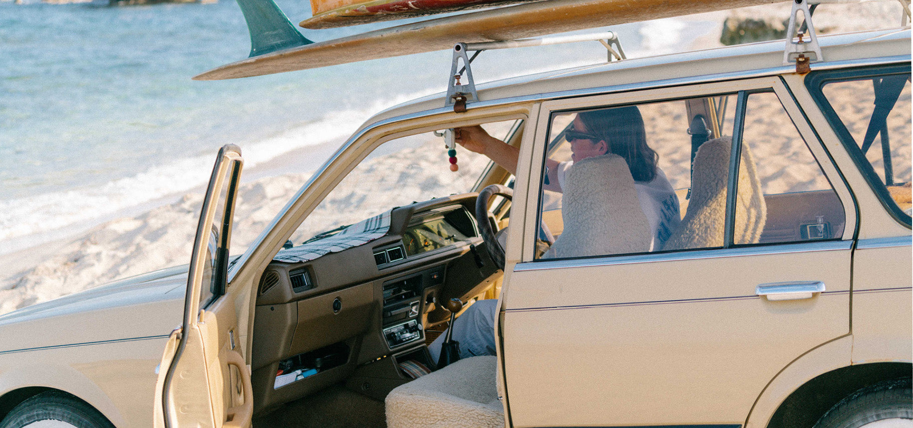 A man sitting in the driver's seat of and old holden commodore hanging up his Smelly Balls reusable air freshener on the beach with surfboards on his roof racks 